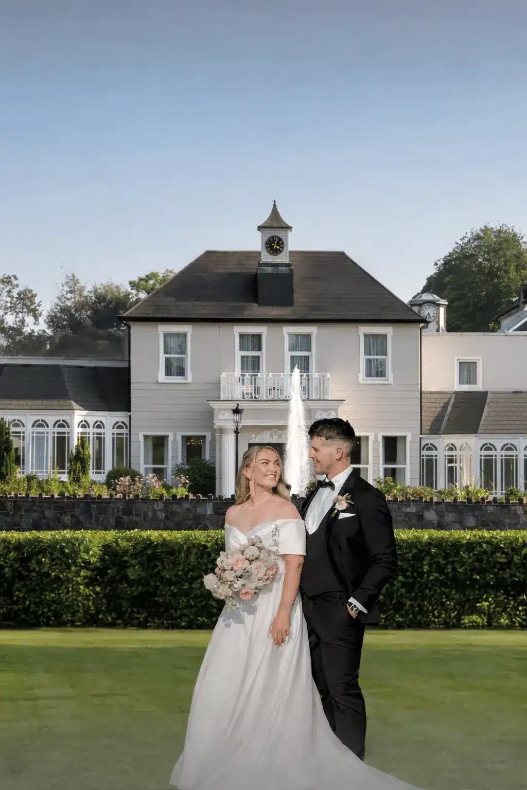 Bride and groom standing in front of a wedding venue with a fountain.